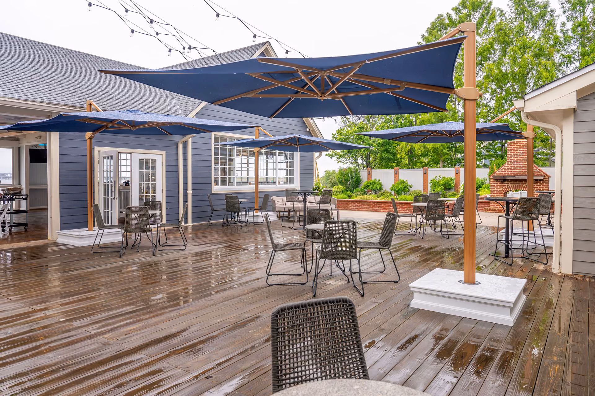 Outdoor patio with wet wooden deck, metal tables and chairs, large blue umbrellas, and string lights overhead.
