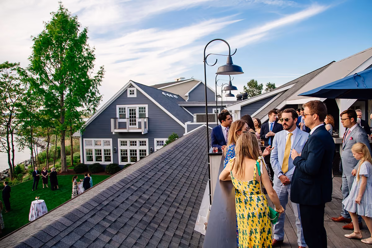 People in formal attire socialize on a rooftop deck with a gray house and lawn gathering visible in the background.