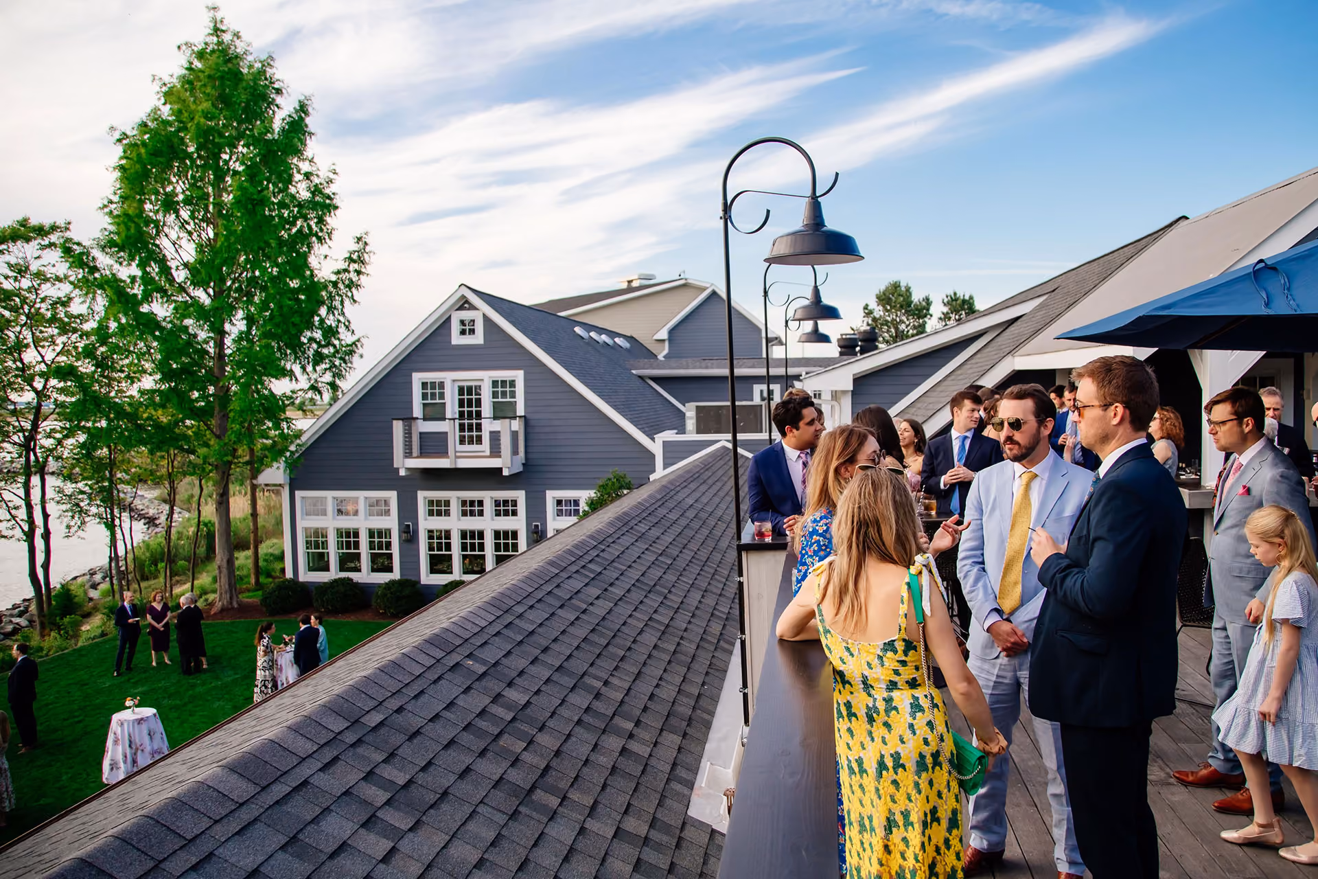 People in formal attire socialize on a rooftop deck with a gray house and lawn gathering visible in the background.