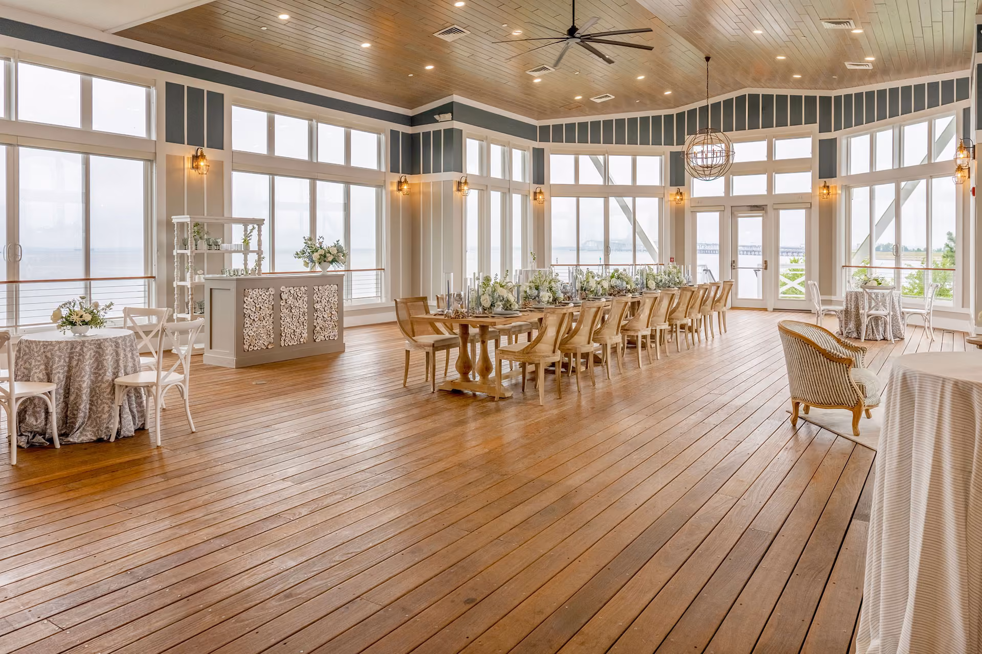 Spacious, bright event hall with wooden floors, large windows, decorated tables, and a bar area.