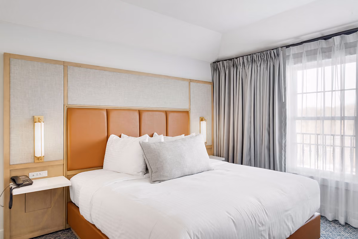 Modern hotel room with a neatly made bed, beige headboard, bedside table, phone, and large window with curtains.