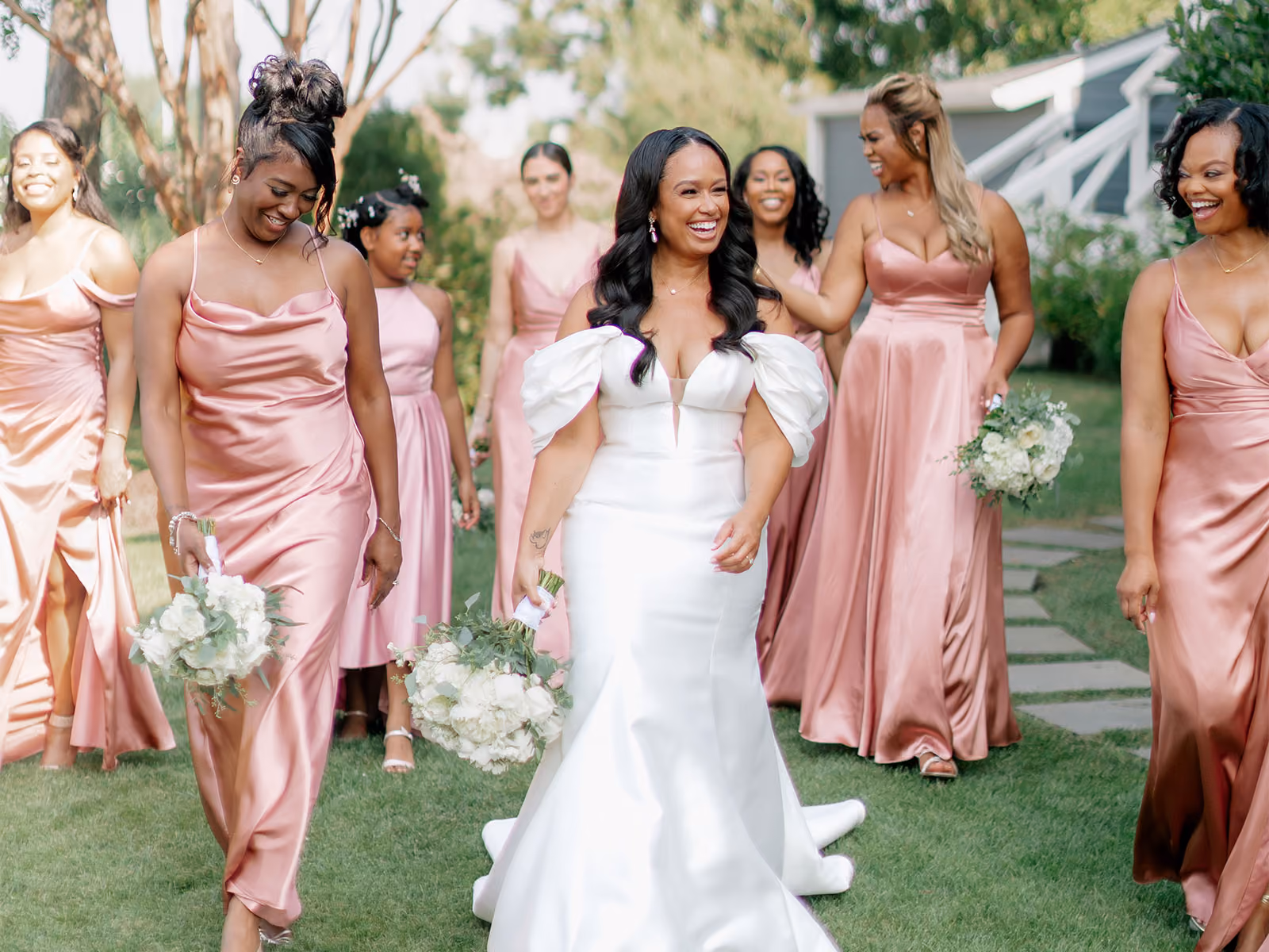 Smiling bride in a white gown walking outdoors with her bridesmaids in rose-pink dresses holding bouquets
