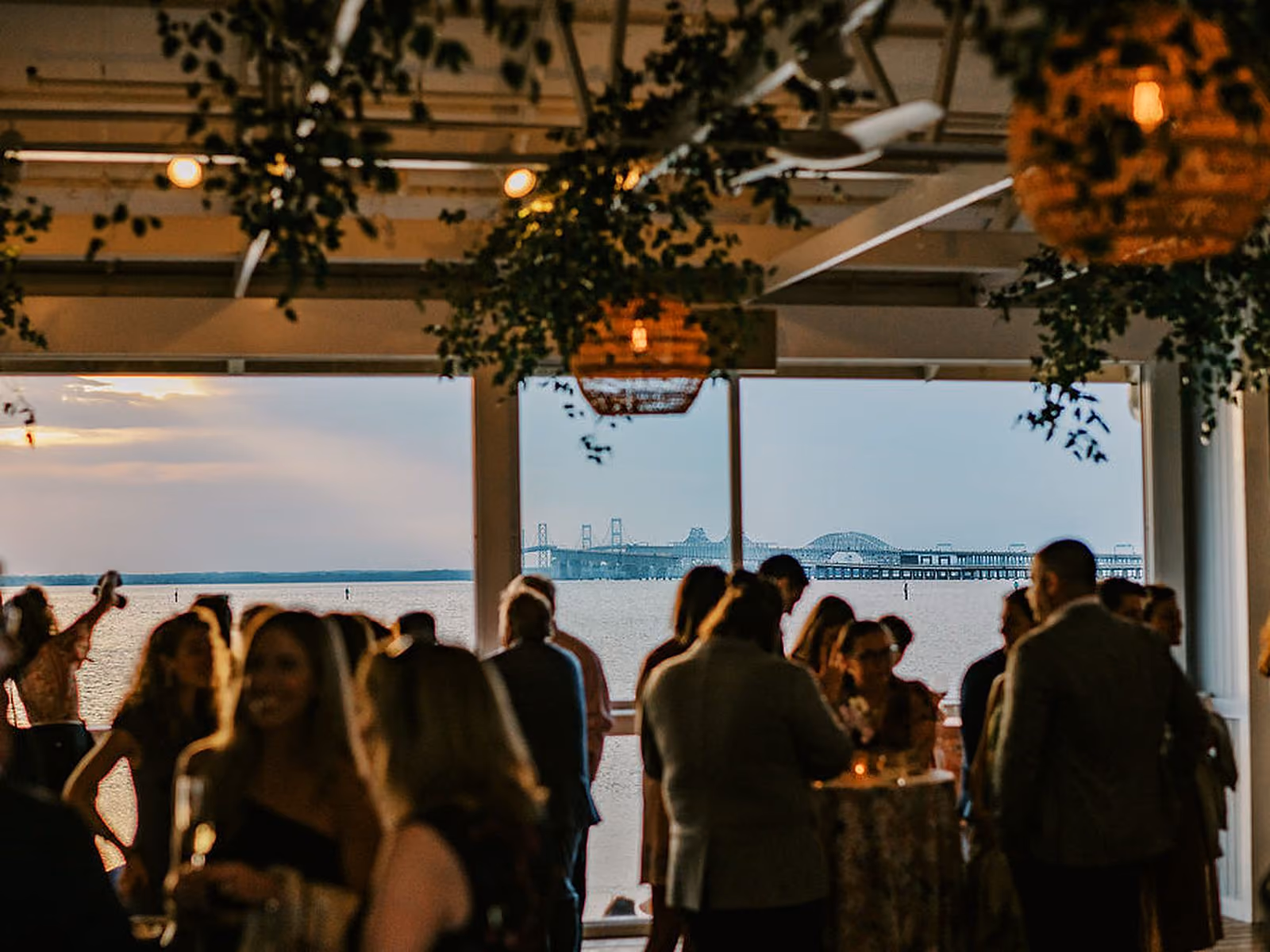 Guests at a waterfront wedding reception with the Chesapeake Bay Bridge framed by windows at sunset