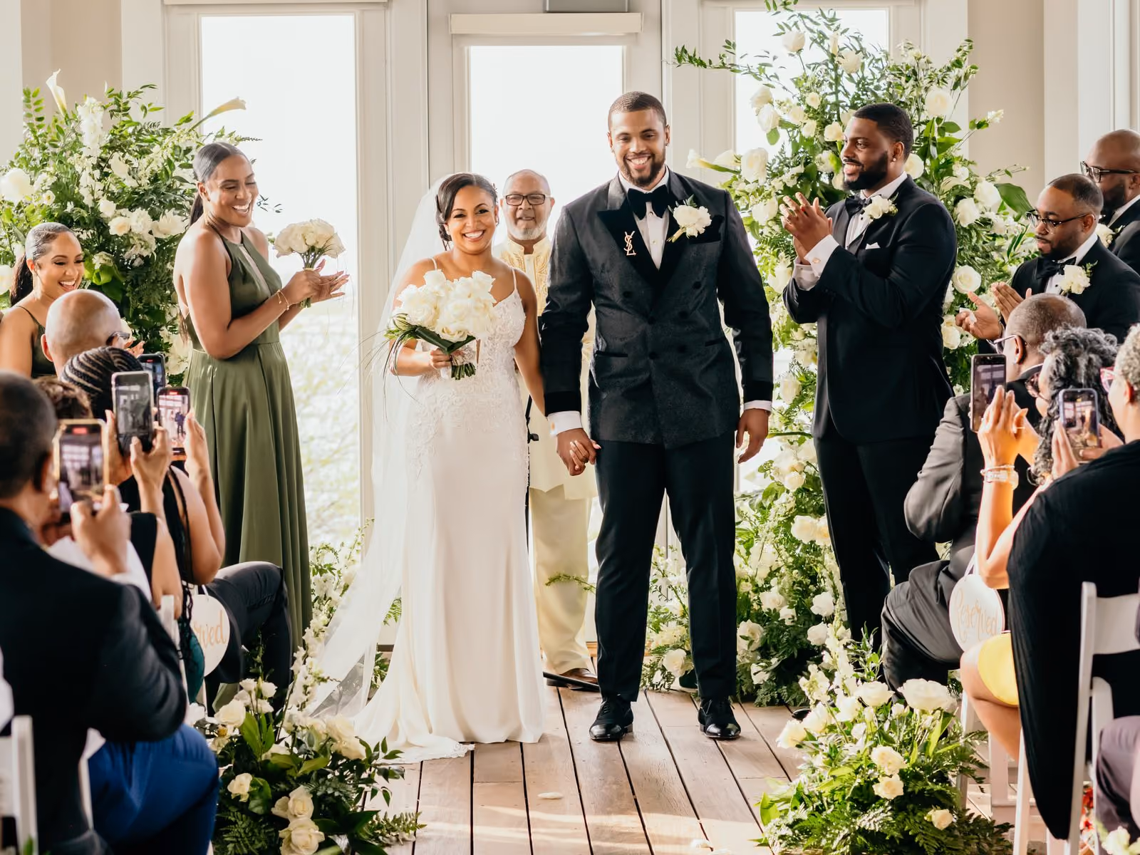 Newlywed bride and groom walking the aisle surrounded by white florals as guests applaud at their ceremony