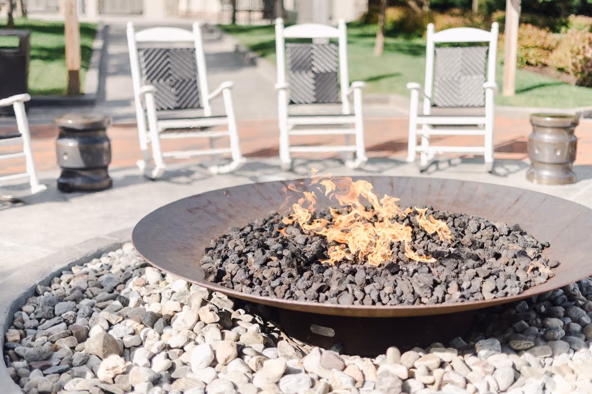 Outdoor fire pit with flames over lava rock and white rocking chairs in the background on a sunny day