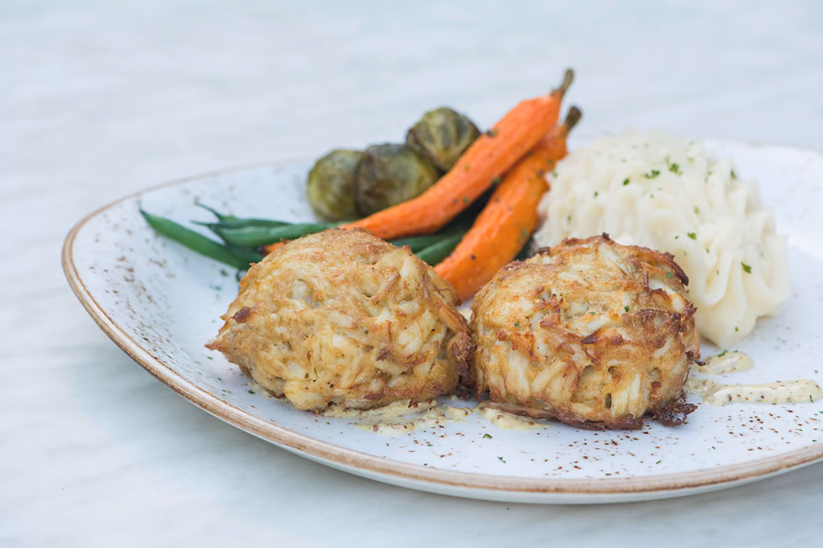 Plated Maryland crab cakes with mashed potatoes, roasted carrots and Brussels sprouts on a speckled plate