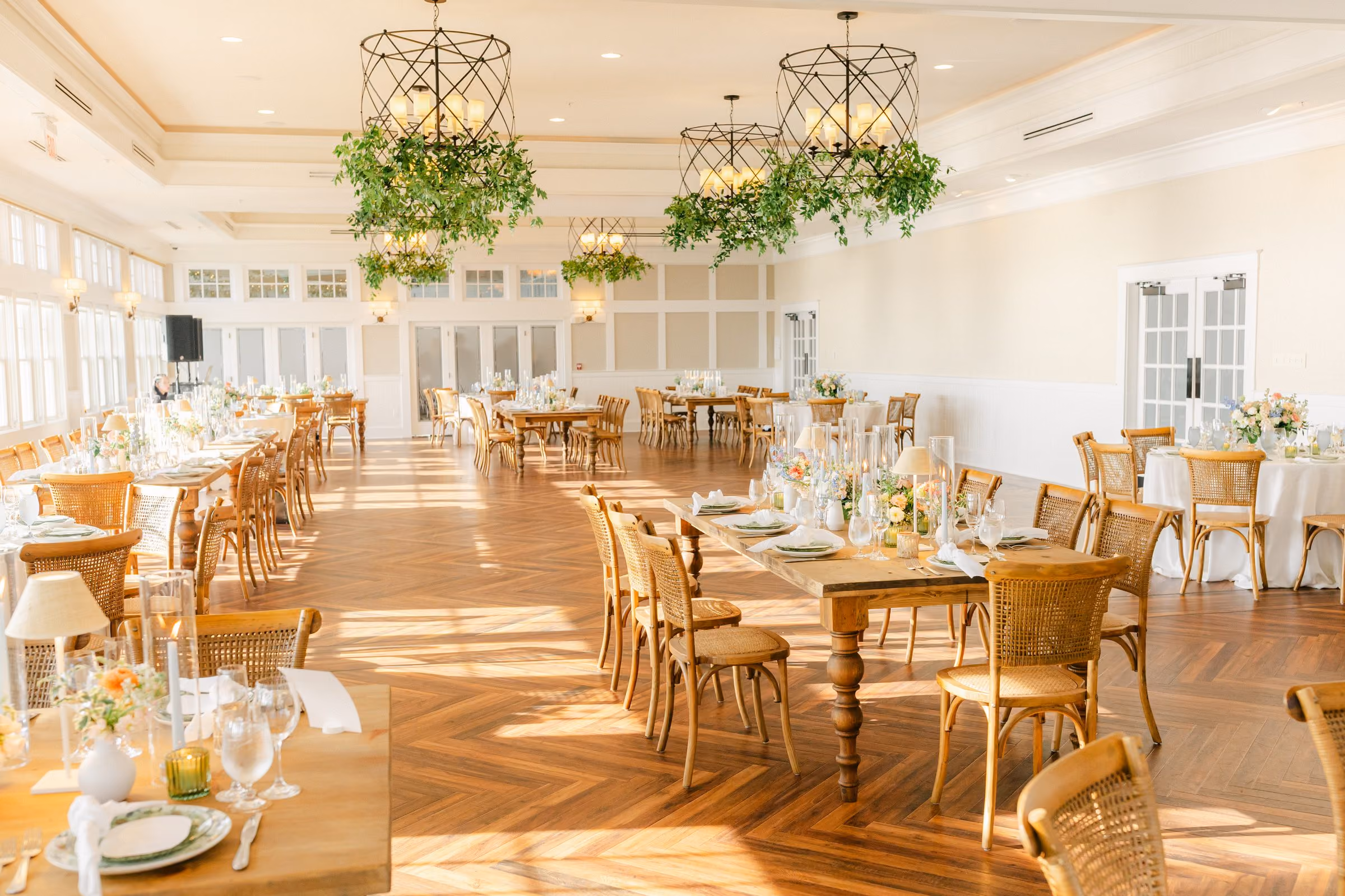 Sunset Ballroom set for a wedding reception with herringbone wood floors, greenery chandeliers and farm tables at Chesapeake Bay Beach Club