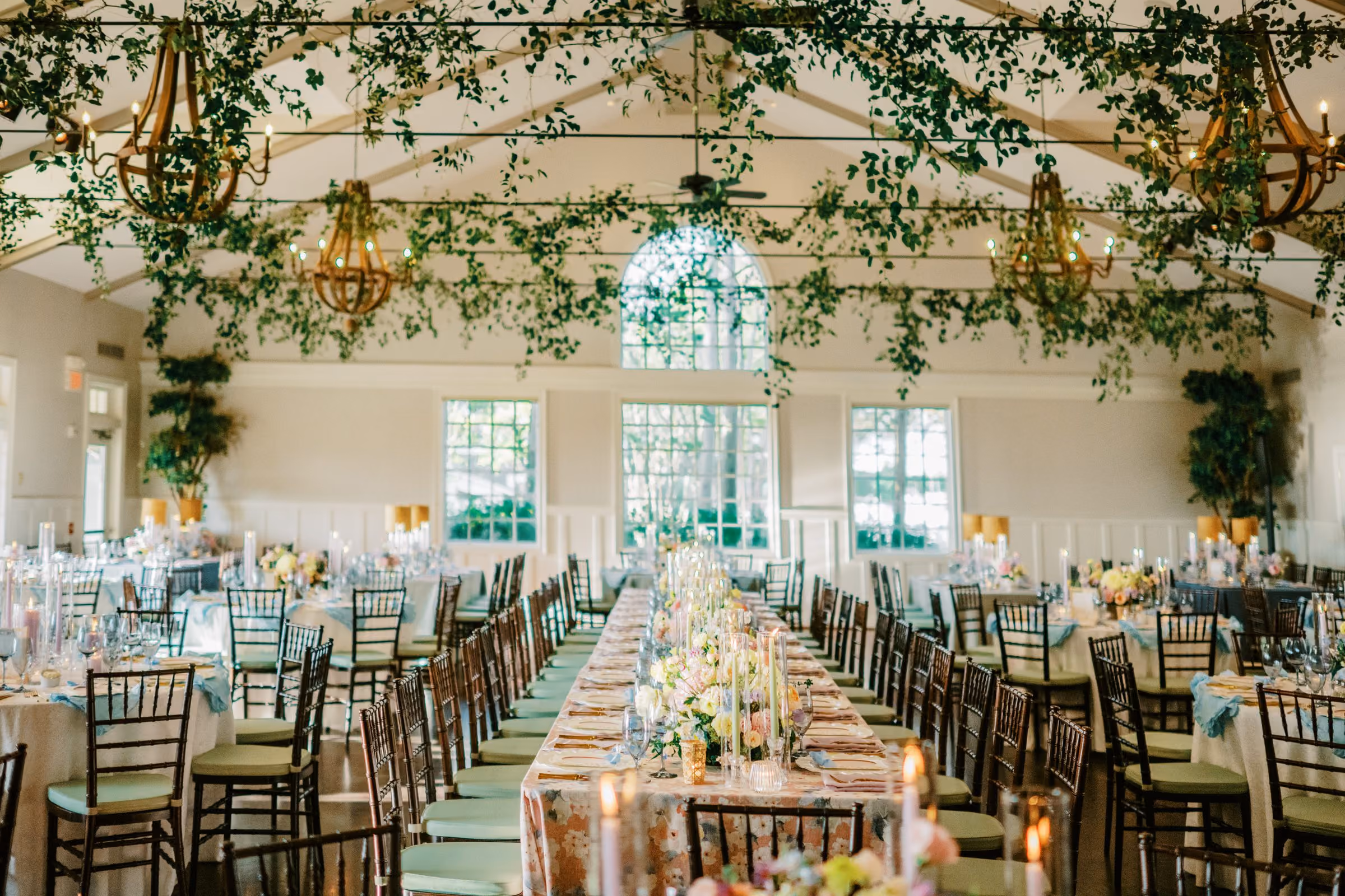 Beach House wedding reception hall with hanging greenery, wrought-iron chandeliers and long banquet tables at Chesapeake Bay Beach Club