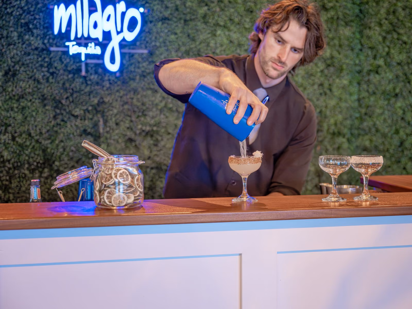 Bartender pouring a craft cocktail at an event bar with a greenery hedge wall and neon sign