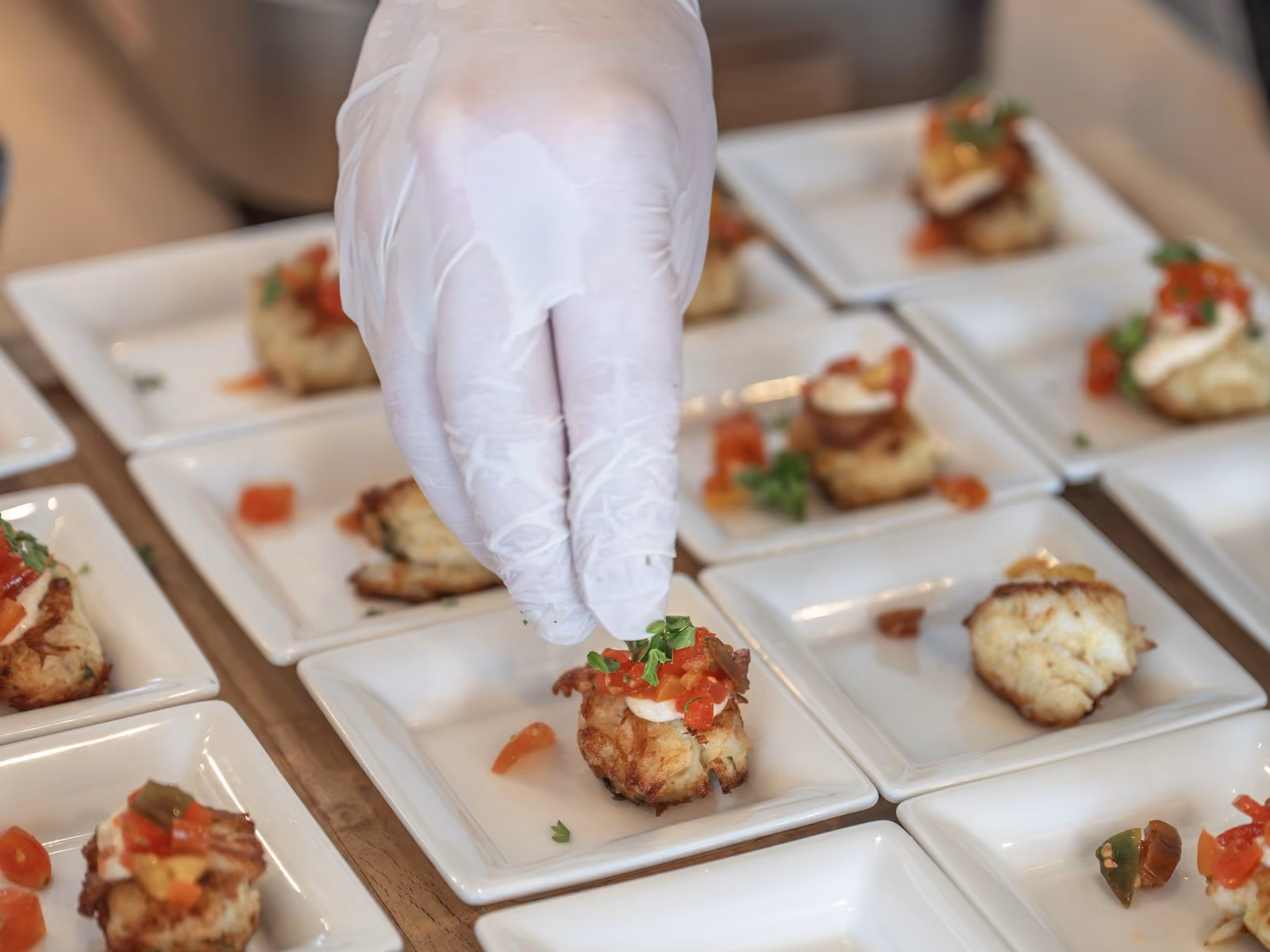Chef plating mini crab cake hors d'oeuvres garnished with diced tomato on white square plates
