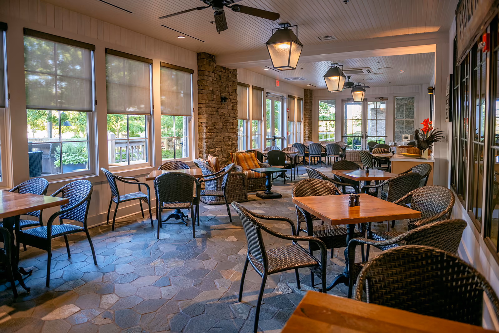 A sunlit indoor cafe seating area with wicker chairs and wooden tables, decorated with plants and vintage signs.