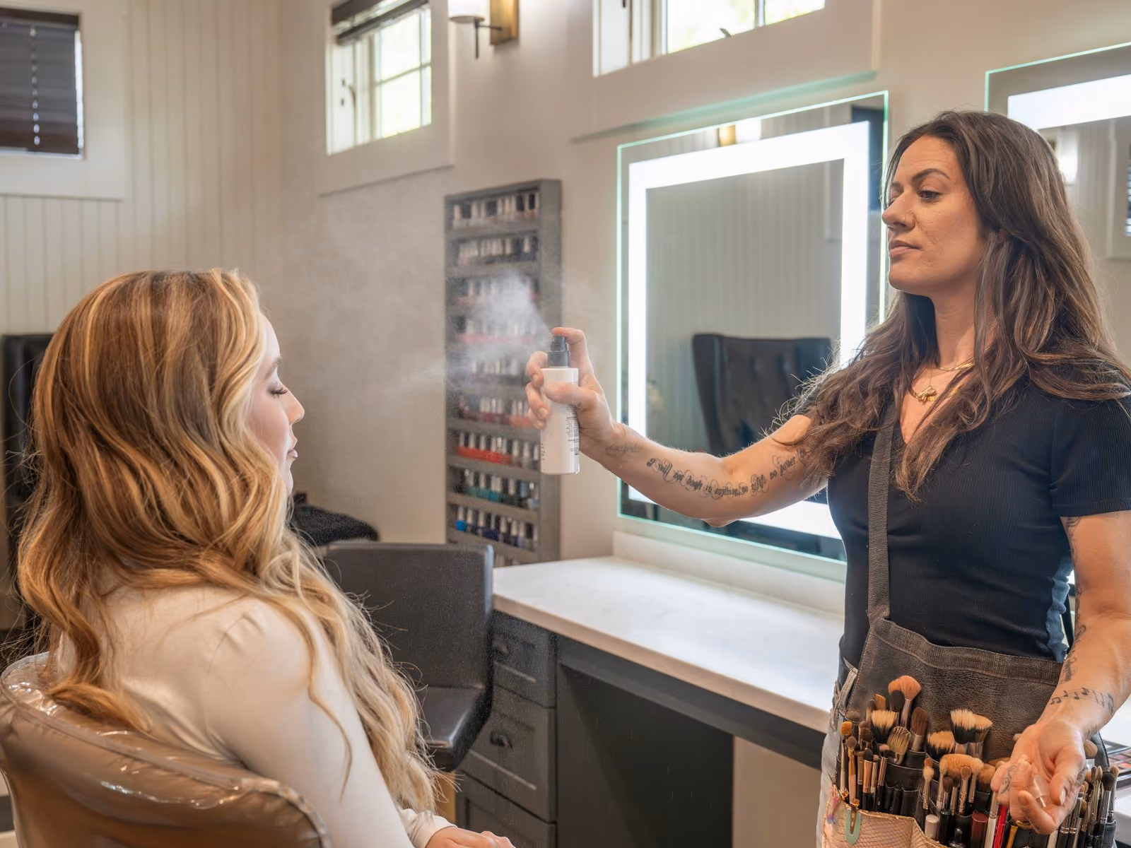 Makeup artist applying setting spray to a bride seated at a lit salon vanity during bridal makeup