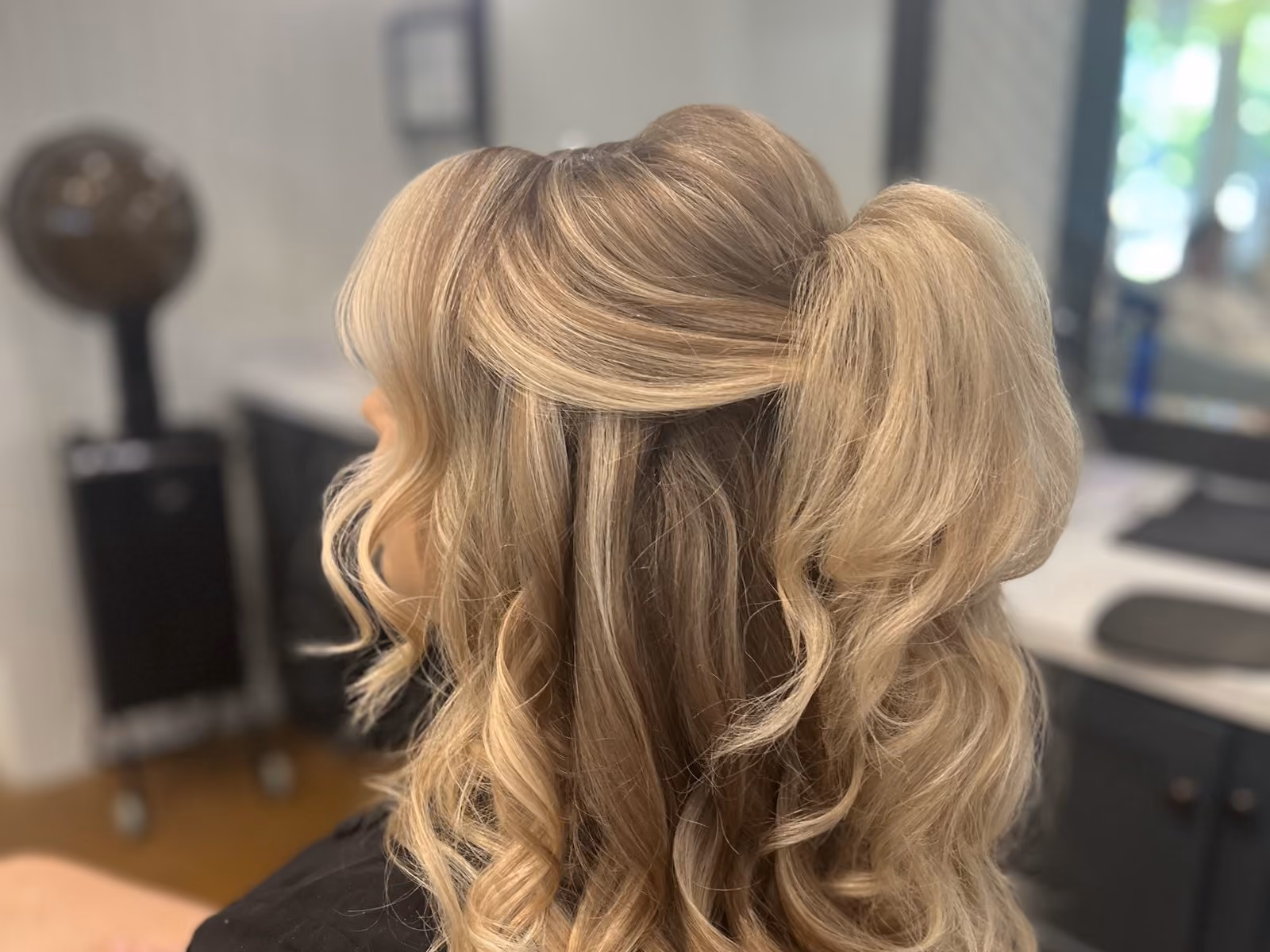 Bride's long curled blonde half-up hairstyle styled for a wedding, viewed from behind in the salon