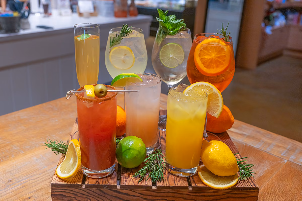 An assortment of colorful cocktails garnished with citrus, olives, and herbs, displayed on a wooden tray.
