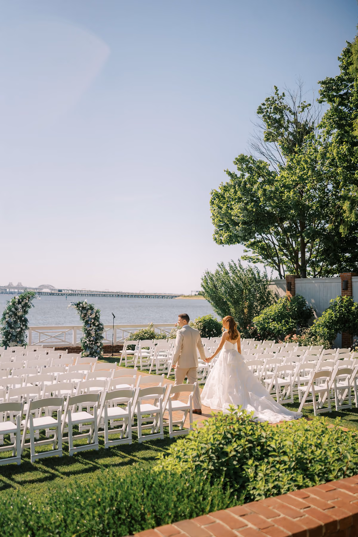A couple in wedding attire walks through rows of white chairs set up outdoors by the water on a sunny day.