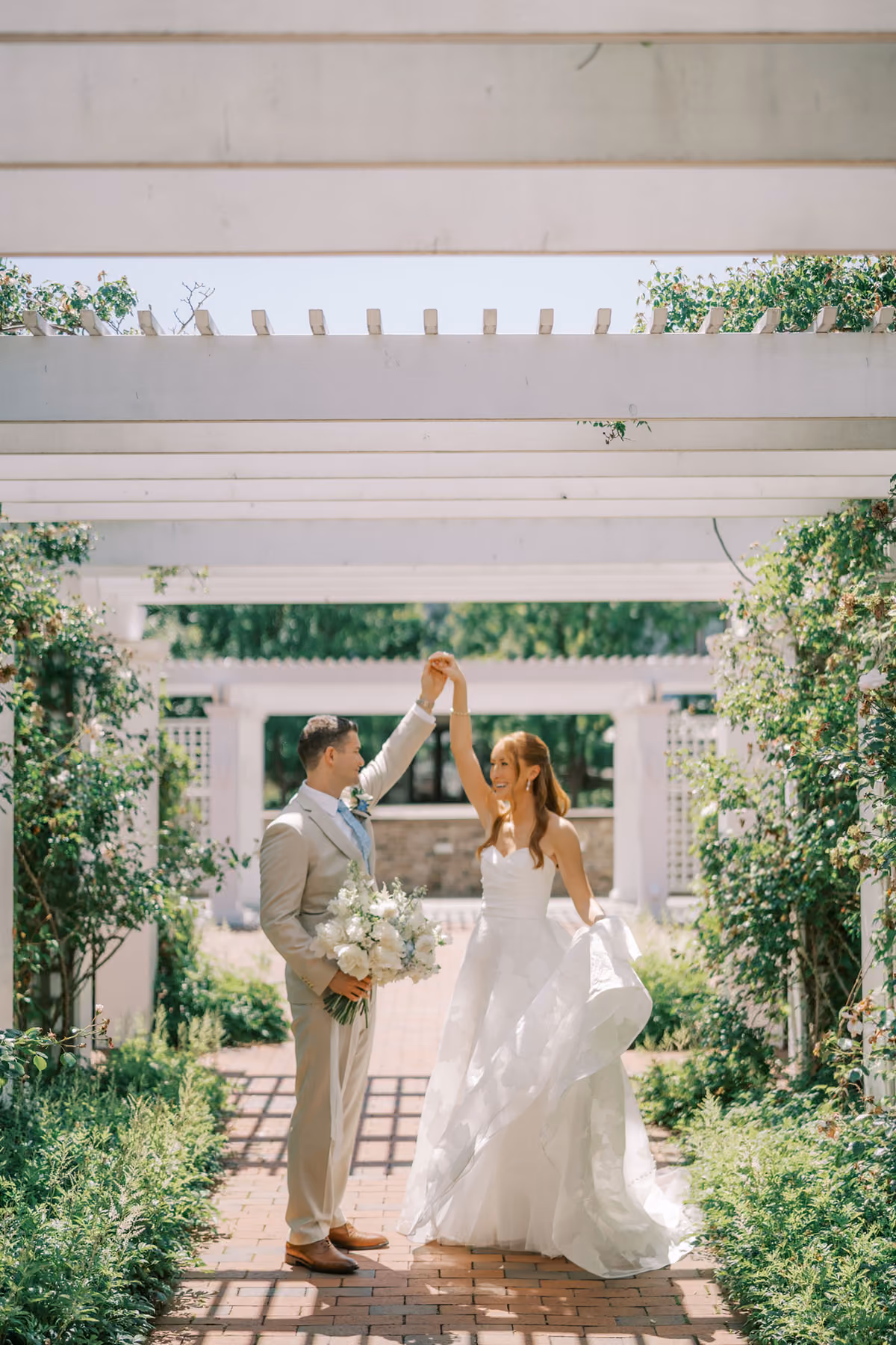 A bride and groom dance together outdoors under a white pergola surrounded by greenery on a sunny day.