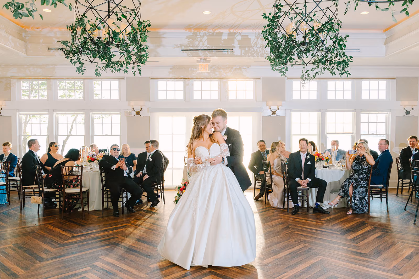 A bride and groom share their first dance in a sunlit reception hall as guests watch from their tables.
