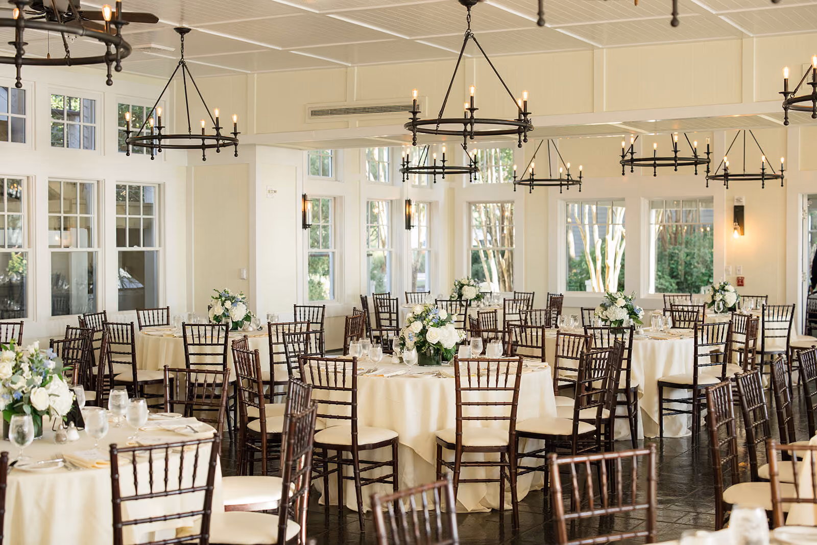 Elegant banquet hall with round tables, dark wooden chairs, floral centerpieces, and large windows letting in natural light.