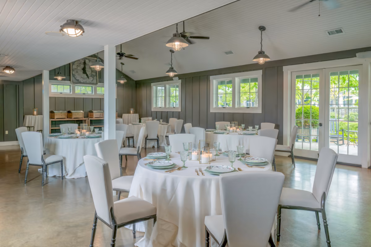 A dining room with round tables, blue chairs, place settings, lantern centerpieces, and overhead lights.