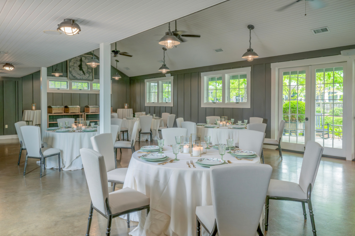 A dining room with round tables, blue chairs, place settings, lantern centerpieces, and overhead lights.