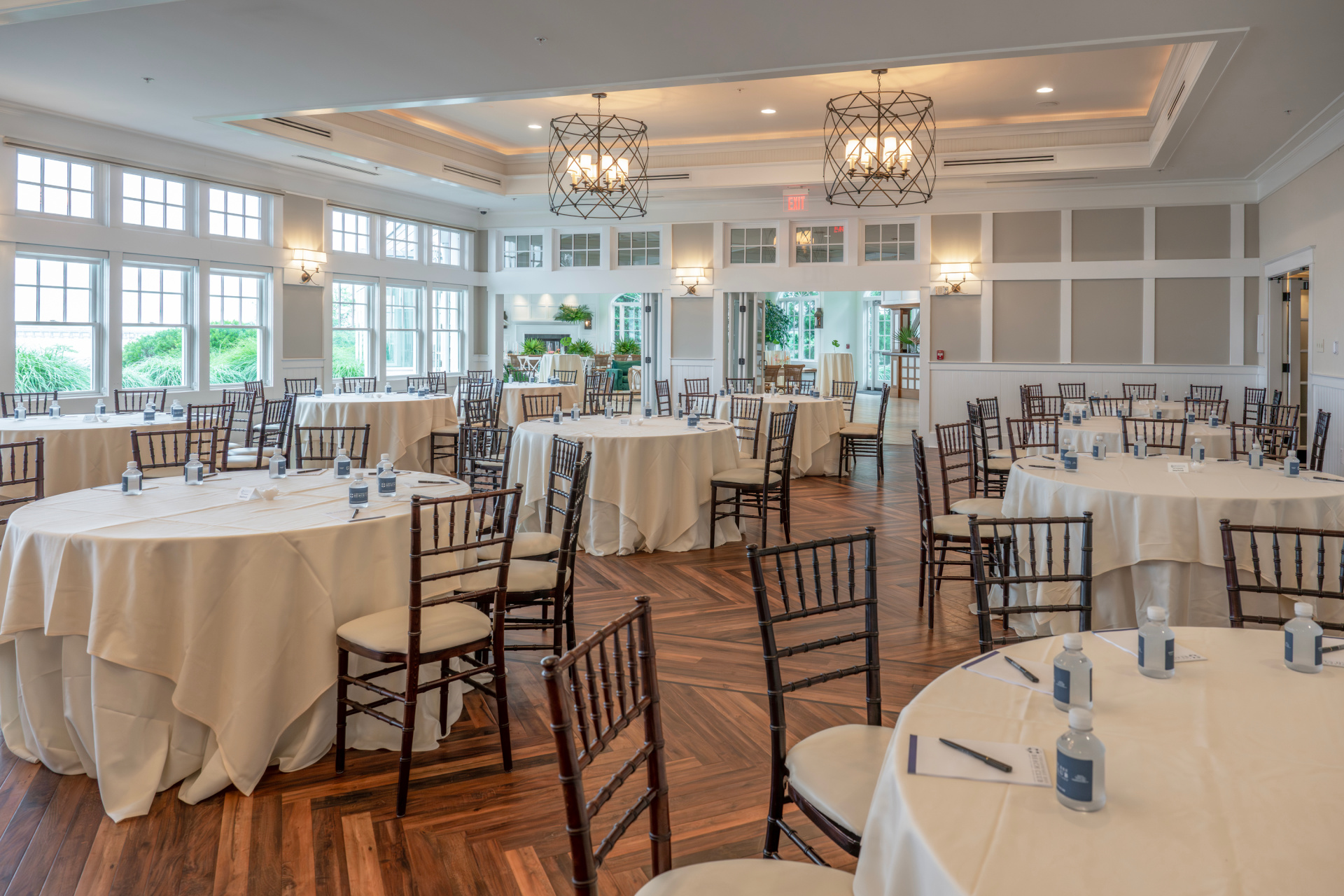 Spacious, bright event hall with wooden floors, large windows, decorated tables, and a bar area.