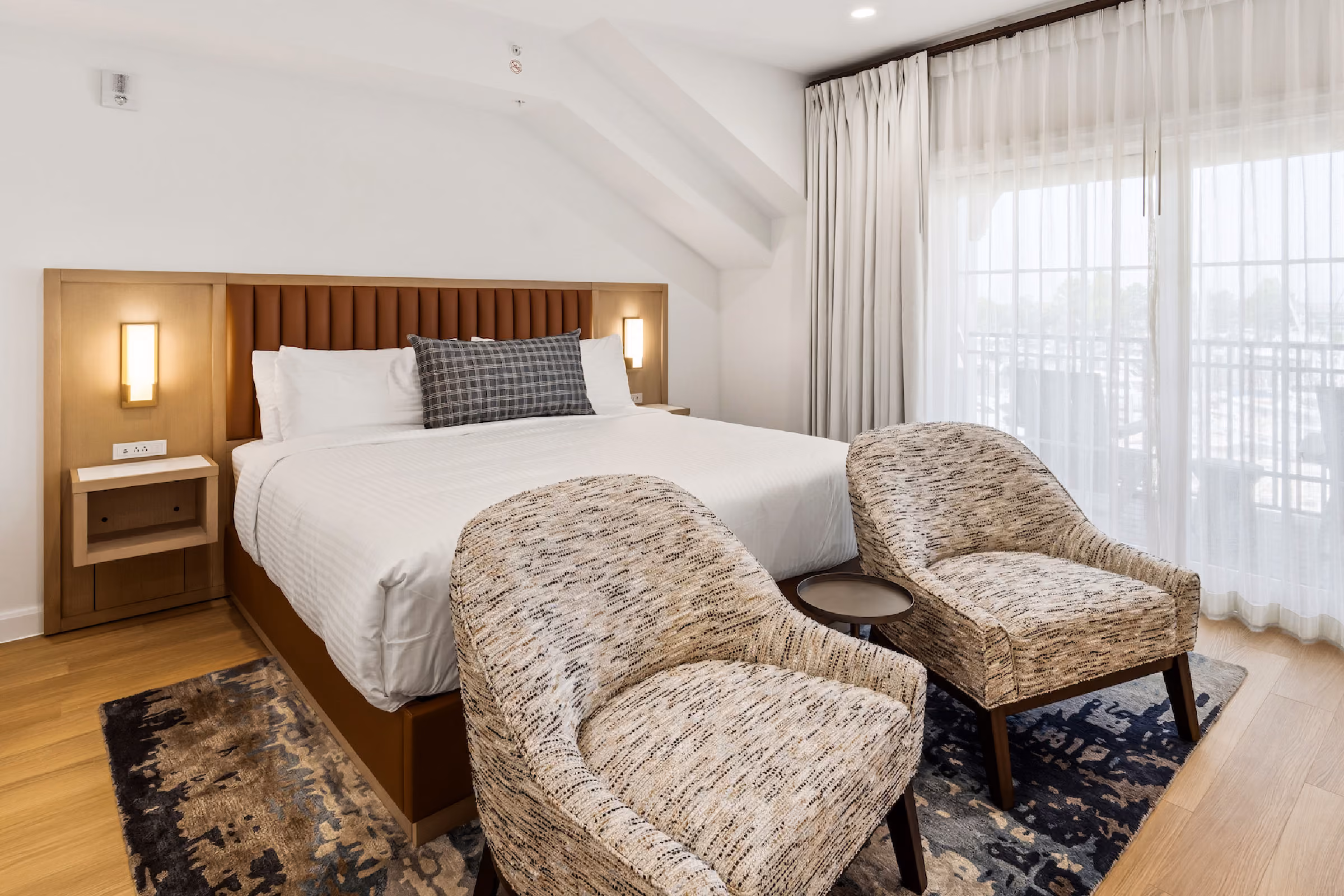 Waterfront hotel room with a large bed, two chairs, desk, and balcony overlooking a sunset view through sliding glass doors.
