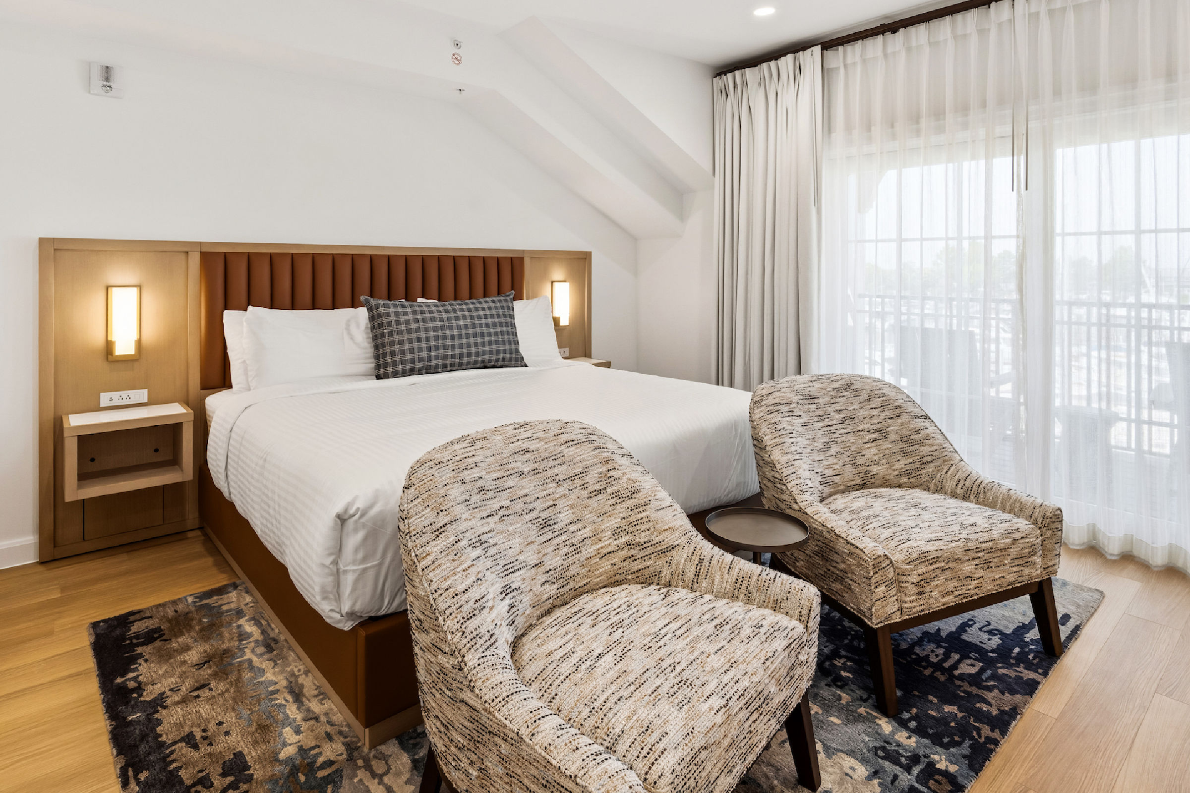 Waterfront hotel room with a large bed, two chairs, desk, and balcony overlooking a sunset view through sliding glass doors.