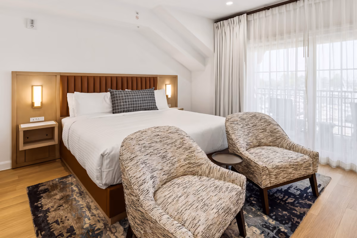 Waterfront hotel room with a large bed, two chairs, desk, and balcony overlooking a sunset view through sliding glass doors.