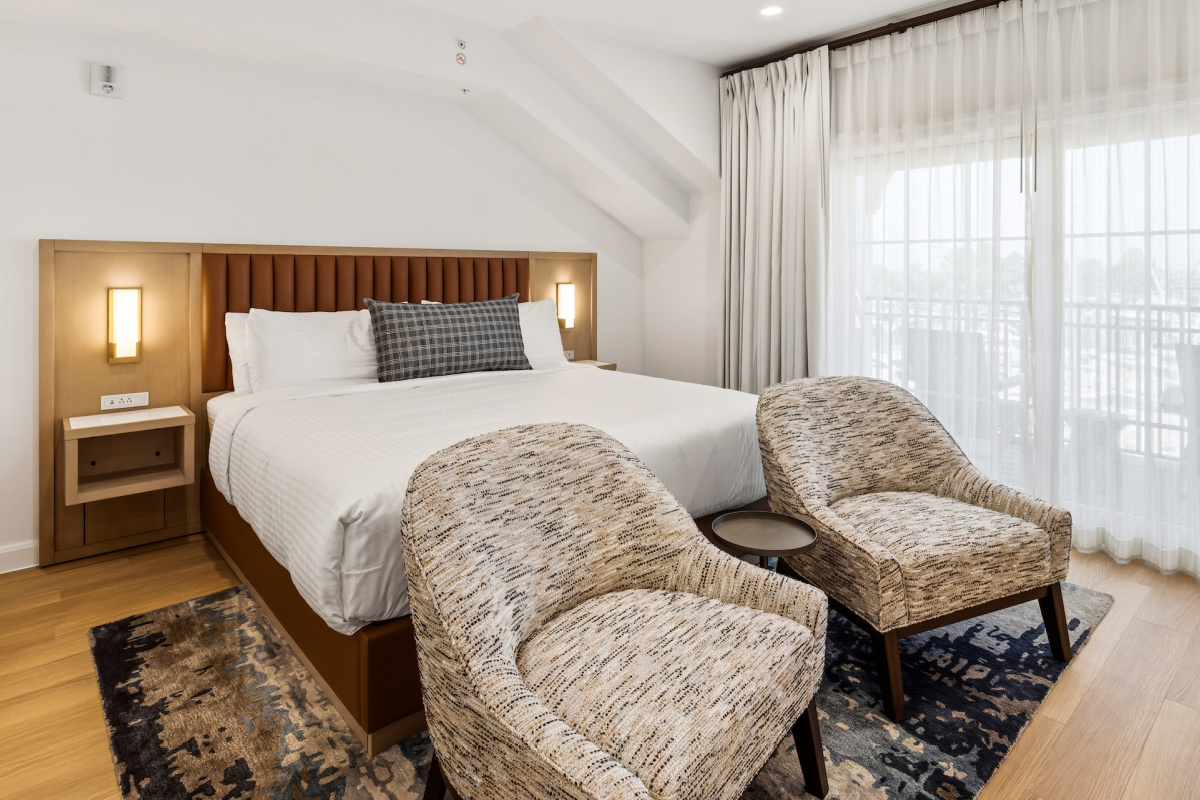 Waterfront hotel room with a large bed, two chairs, desk, and balcony overlooking a sunset view through sliding glass doors.