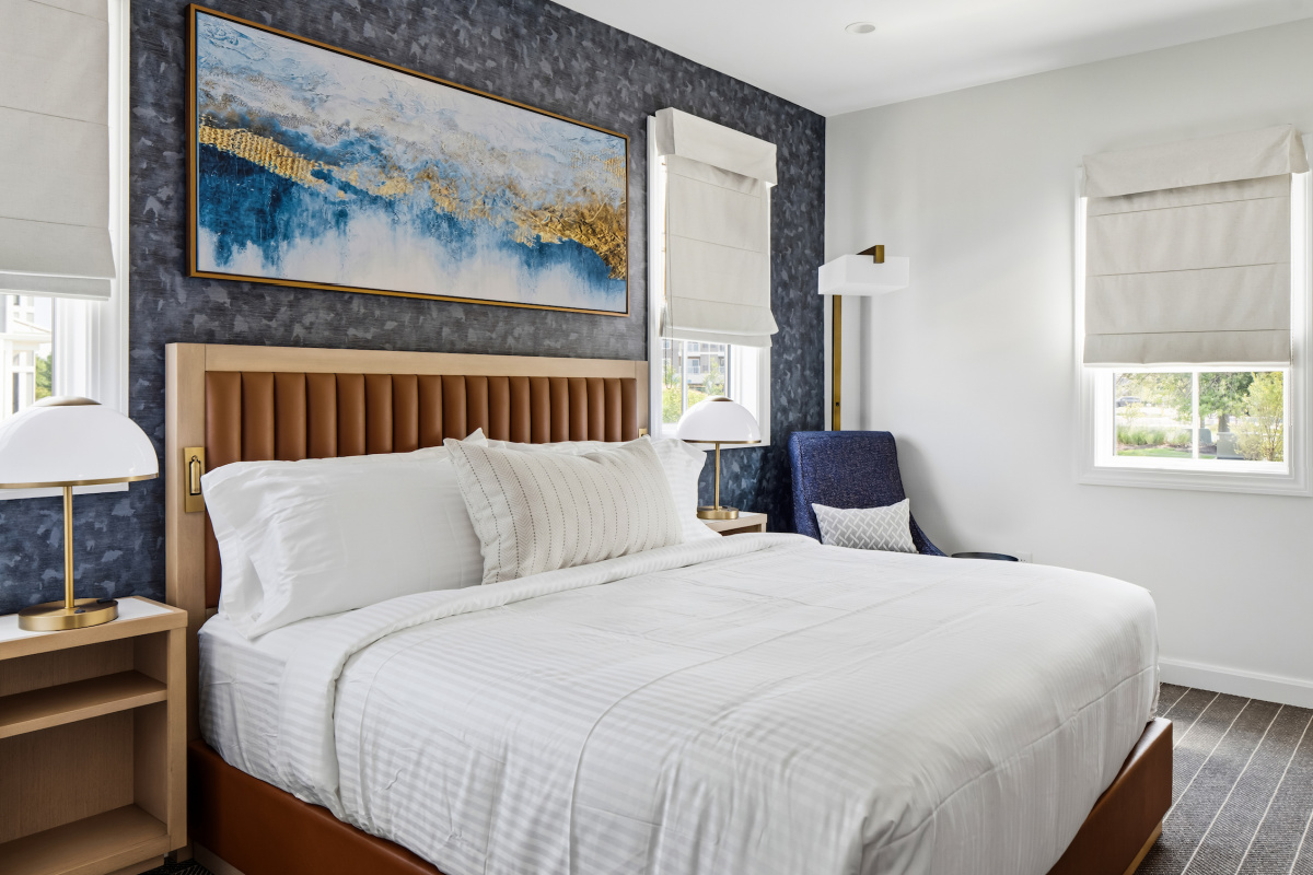 Modern hotel room with a large bed, two chairs, desk, and balcony overlooking a sunset view through sliding glass doors.