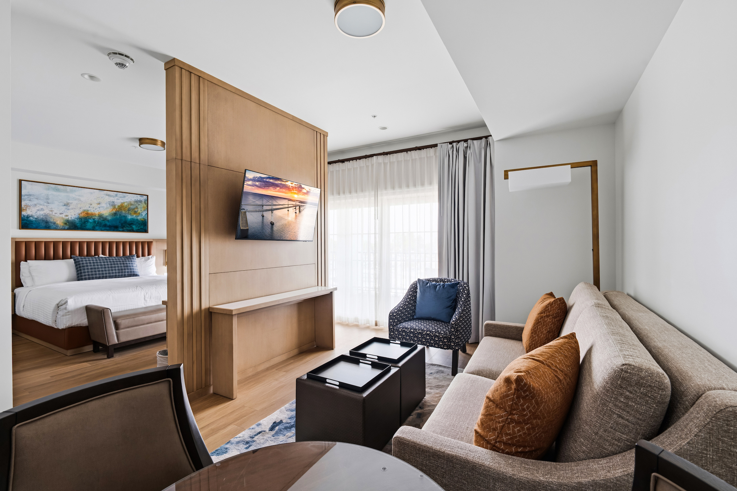 Modern hotel room with a large bed, two chairs, desk, and balcony overlooking a sunset view through sliding glass doors.