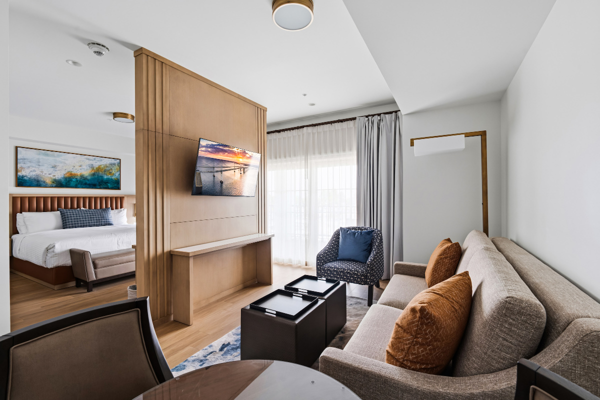Modern hotel room with a large bed, two chairs, desk, and balcony overlooking a sunset view through sliding glass doors.