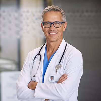 Smiling middle-aged male doctor in white coat with stethoscope and glasses, standing with arms crossed indoors.