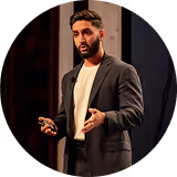 Man with beard and dressed in a dark blazer speaking and gesturing during a presentation on stage.