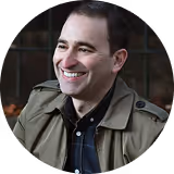 Smiling man with short dark hair wearing a green jacket over a dark shirt.