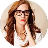 Woman with long reddish hair, red lipstick, glasses, and a white scarf against a beige background.