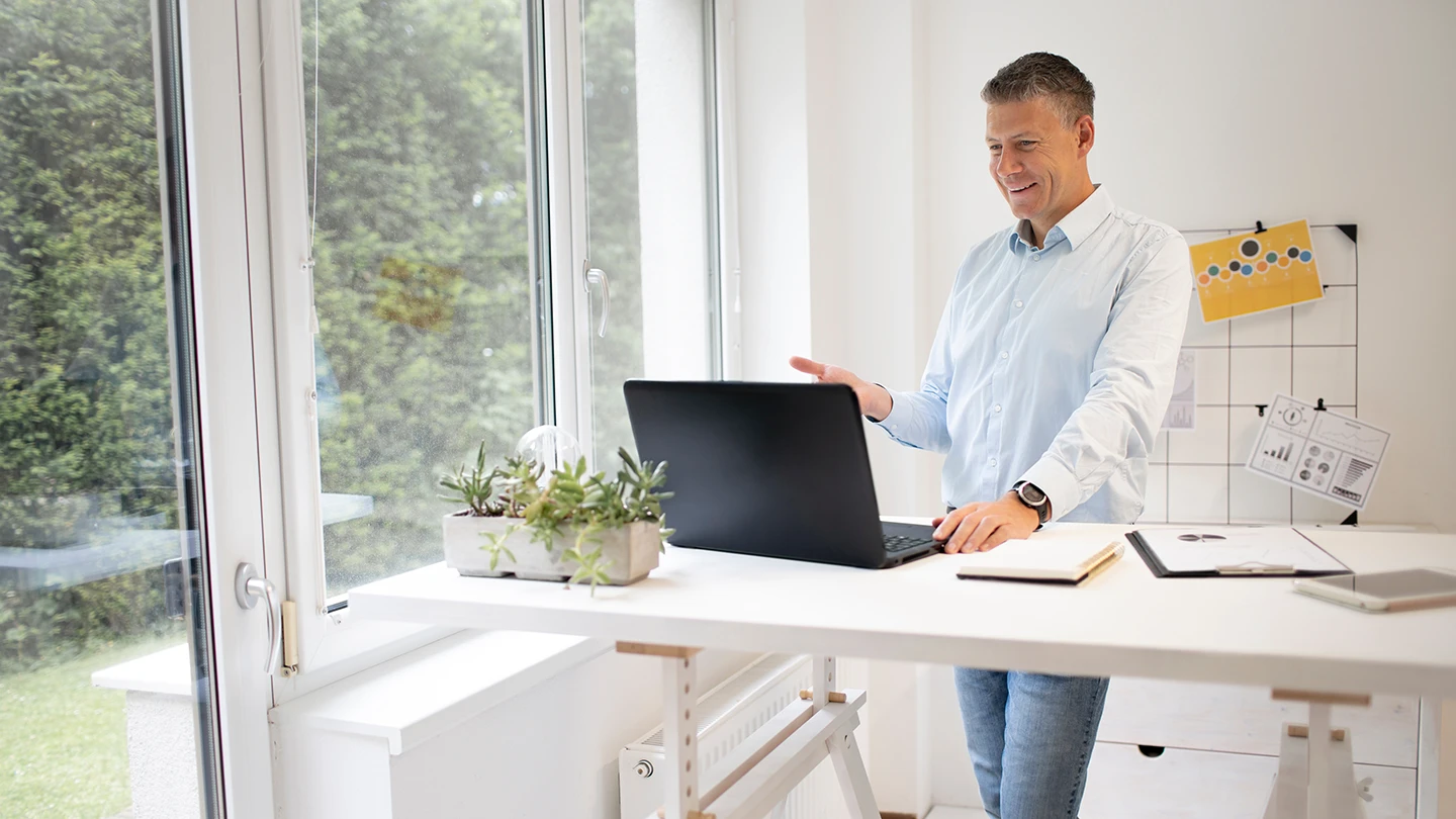 Professional using a height-adjustable standing desk in a commercial office with a laptop, plant, and natural light from a large window