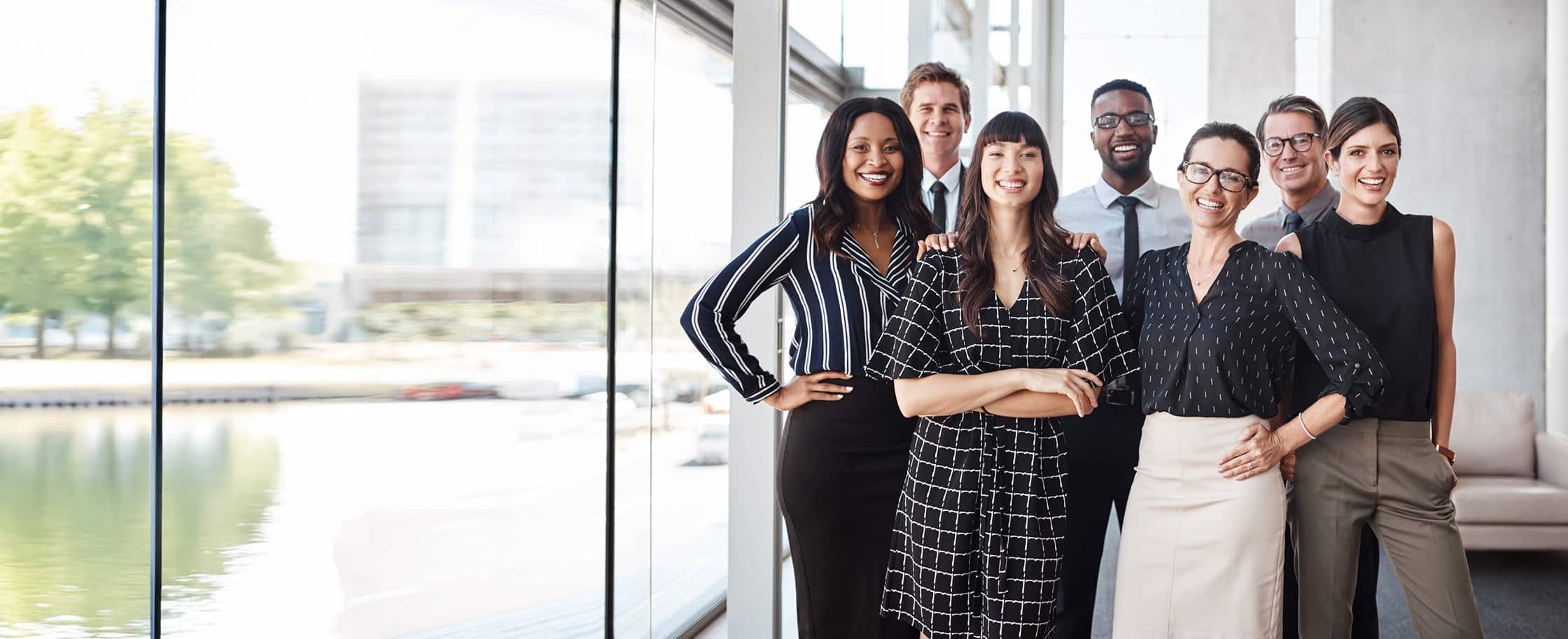 Group of seven diverse business professionals smiling and posing inside a modern office near a window.