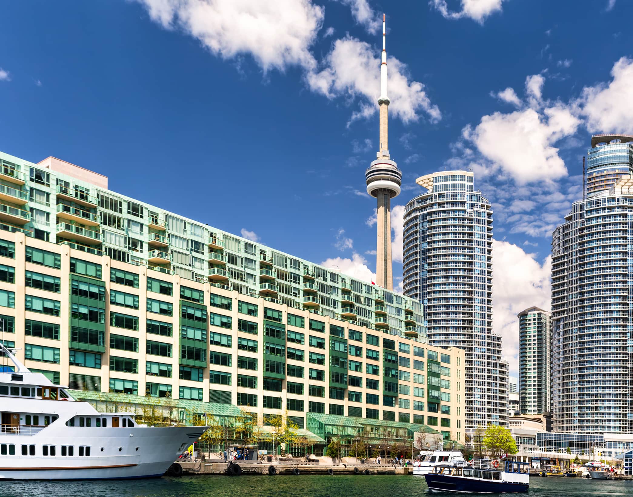 Toronto waterfront with CN Tower, modern high-rise buildings, and boats on water under blue sky.