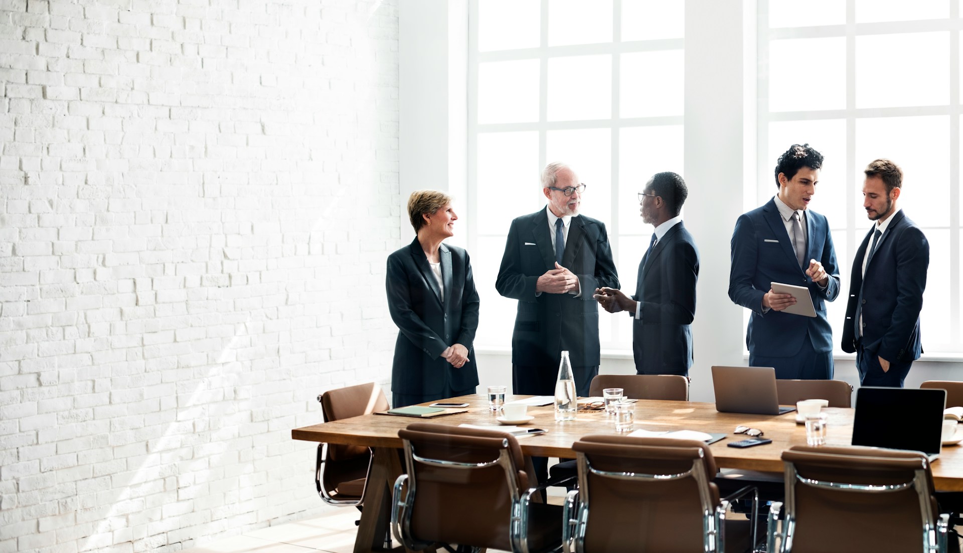 Five businesspeople in suits talking in a bright conference room with table and chairs.
