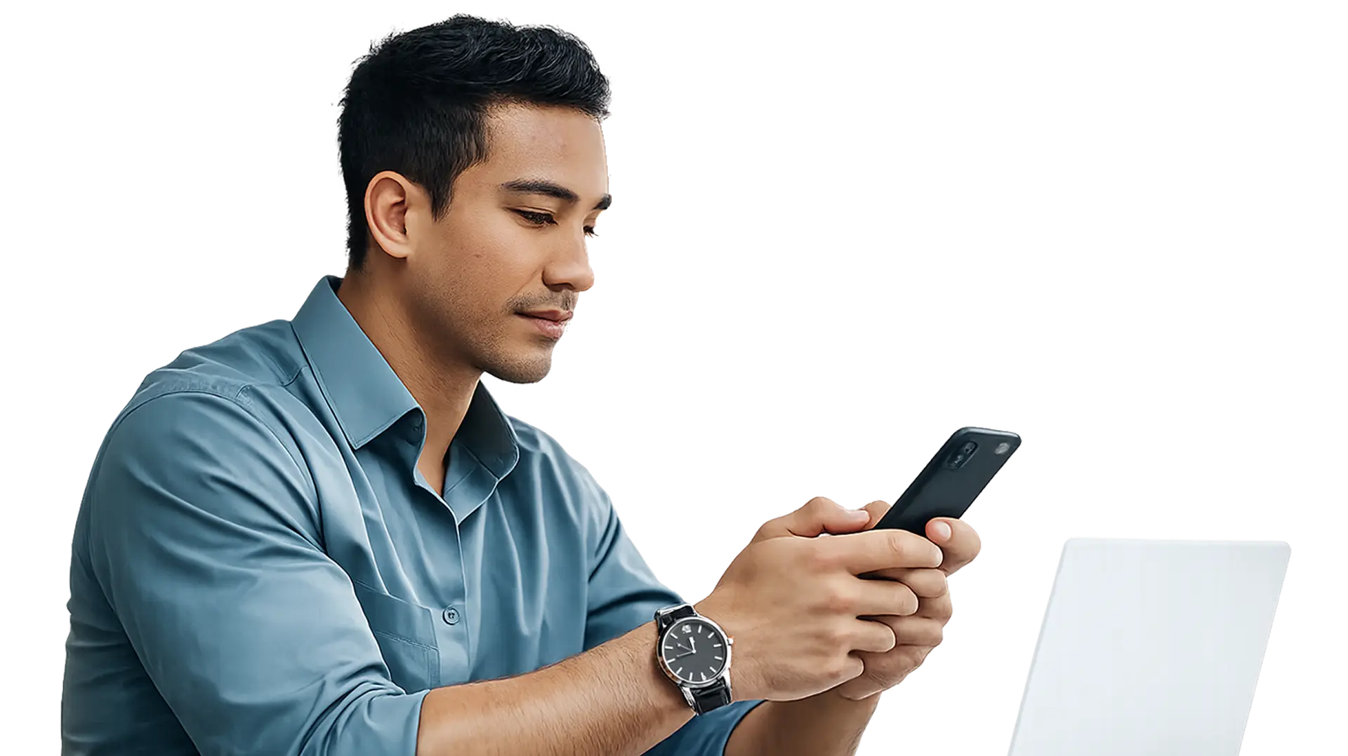 Man in blue shirt holding and looking at a smartphone, with a laptop visible in front of him.