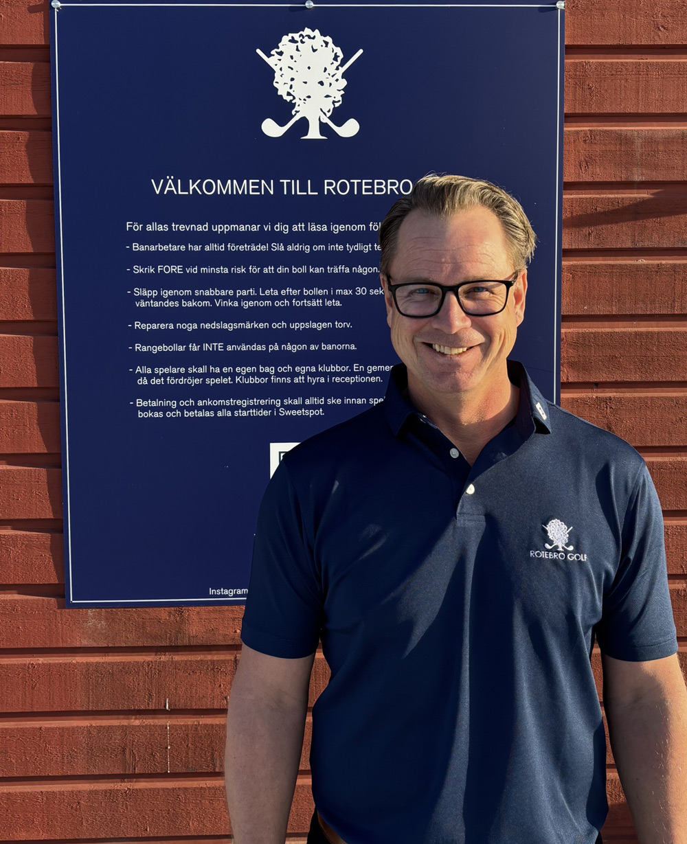 Smiling man wearing glasses and navy blue Rotebro Golf polo standing in front of a wooden wall with a navy sign containing golf rules in Swedish.