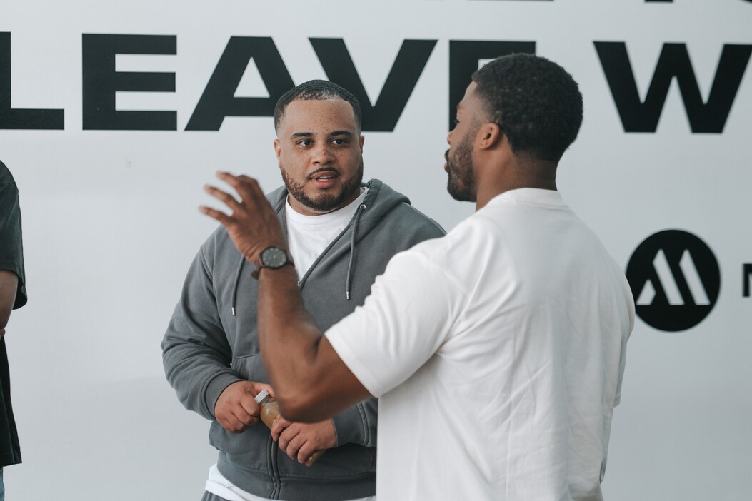 Two men engaged in a conversation in a brightly lit space with partial bold text on the wall behind them.