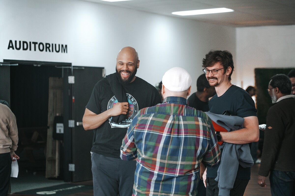 Three men smiling and chatting in a hallway near an auditorium entrance.