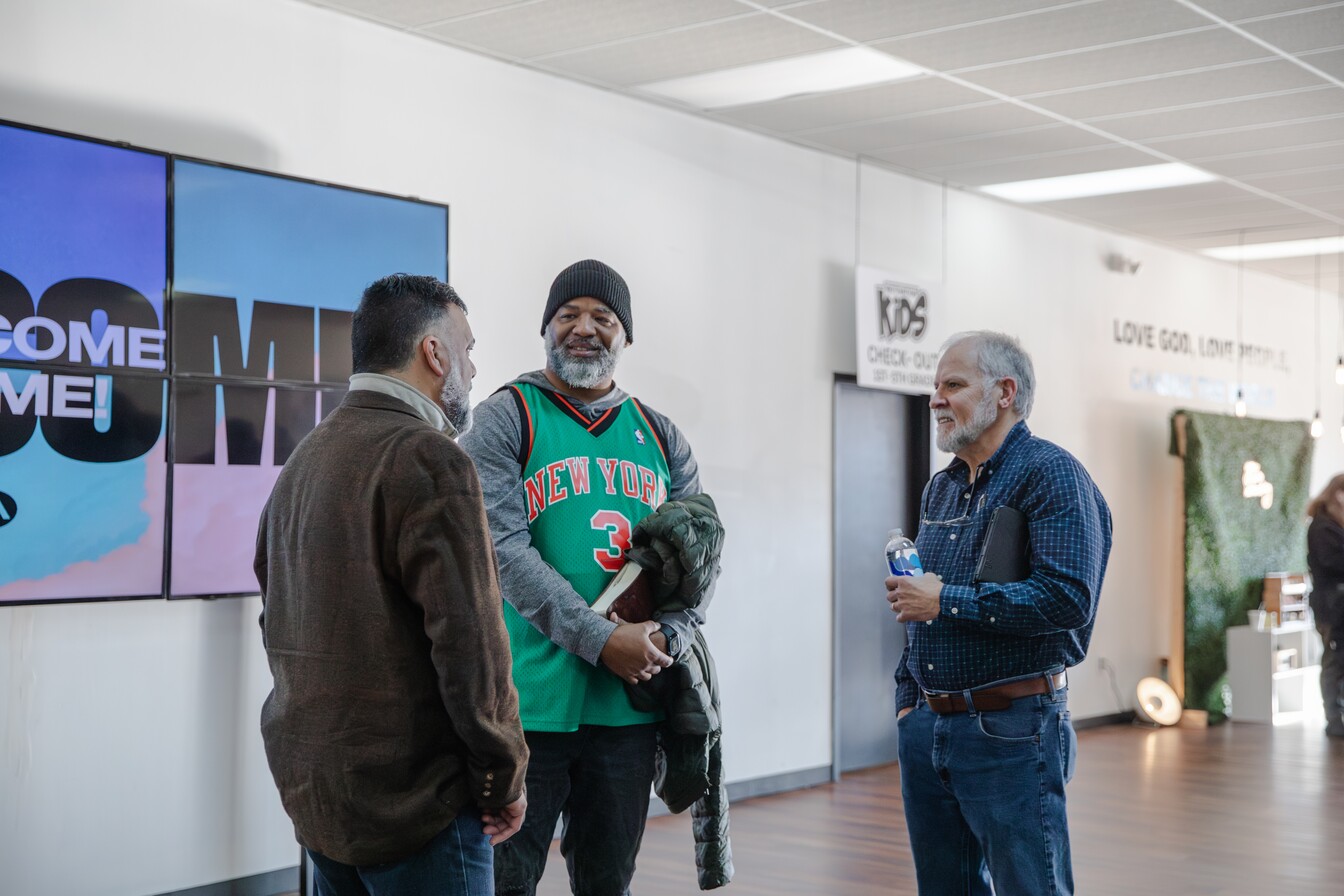 Three men standing indoors, engaged in conversation near a wall with a digital welcome sign and a kids check-in area.