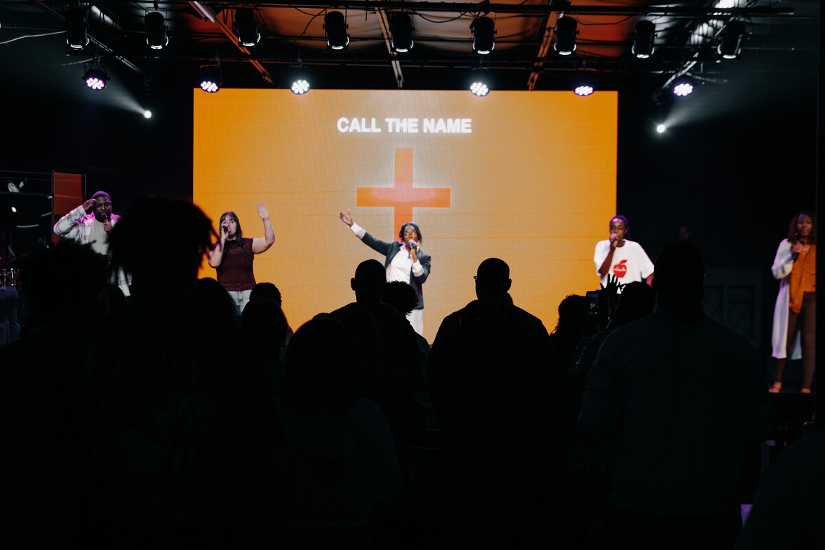 A worship band singing on stage in front of an orange screen with a cross and the words 'CALL THE NAME,' with an audience silhouetted in the foreground.