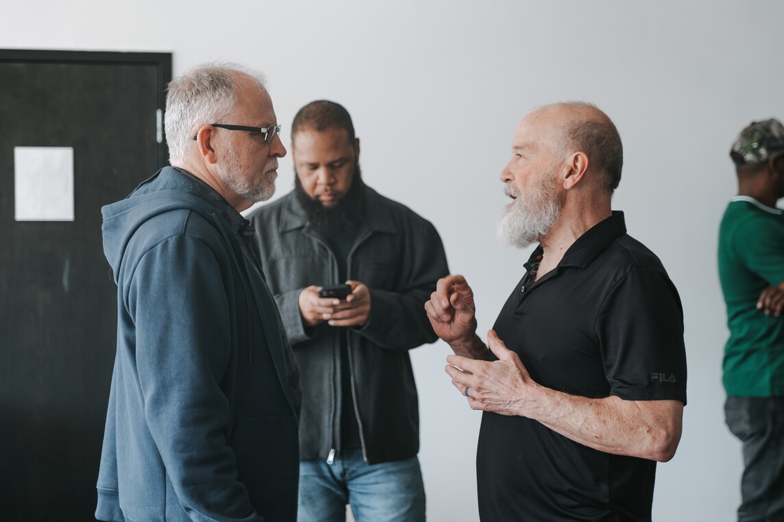 Two older men with gray beards having a conversation indoors while a man behind them looks at his phone and another man in green leans against the wall.