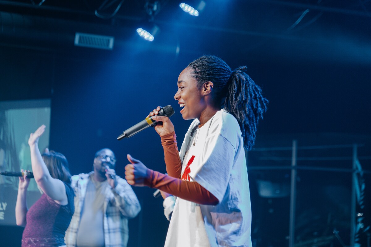 Smiling woman with dreadlocks singing into microphone on stage during a Motivation Church service with two other singers in the background.