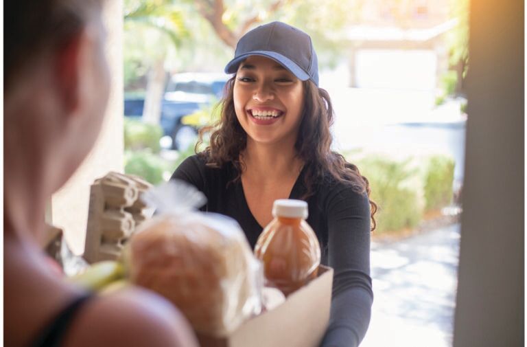 Smiling delivery woman in a blue cap handing a box of groceries including bread and a beverage to a customer at a home doorstep.