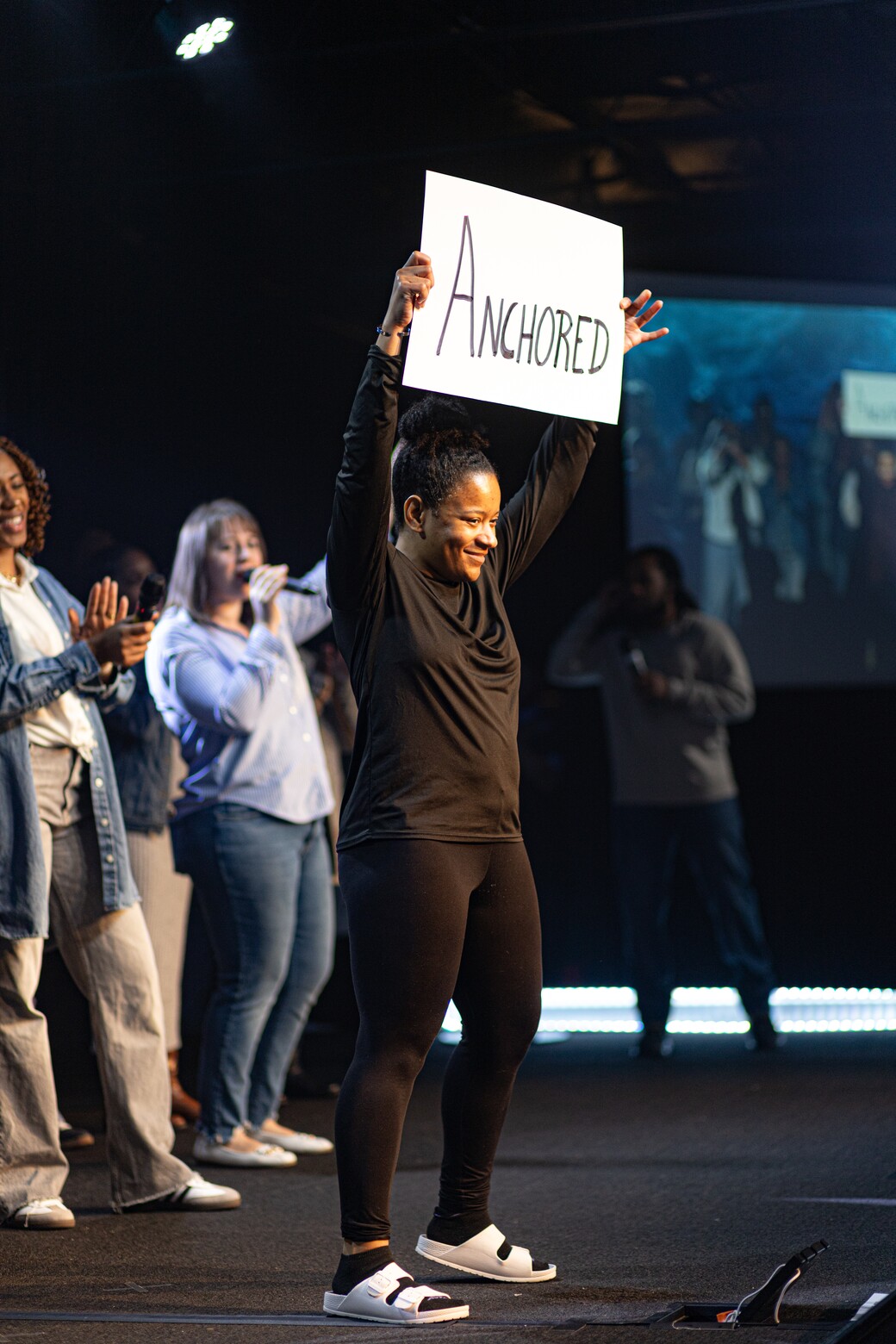 Smiling woman on stage holding a sign reading 'Anchored' above her head with people clapping and singing in the background.
